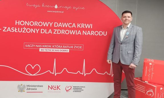 Na zdjęciu Łukasz Kaczyński stojący na tle planszy honorowy dawca krwi